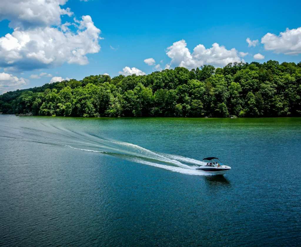 boat swiftly moving across a lake on a partially cloudy sunny day with full green trees on the shore.