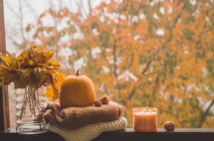 Lit candle, a pumpkin, sweaters, and dead leaves sitting in a window with fall trees outside
