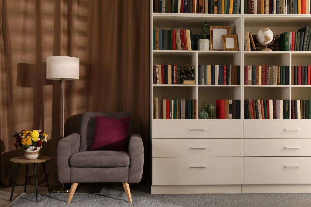 A cozy living room features a white bookshelf filled with books, a brown plush chair, lamp, and side table with flowers
