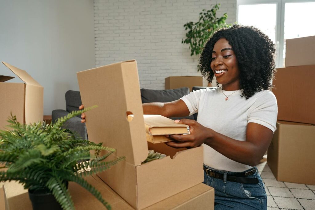 A woman sits on the floor of an apartment packing books into a cardboard box. She is surrounded by other cardboard boxes and packing materials