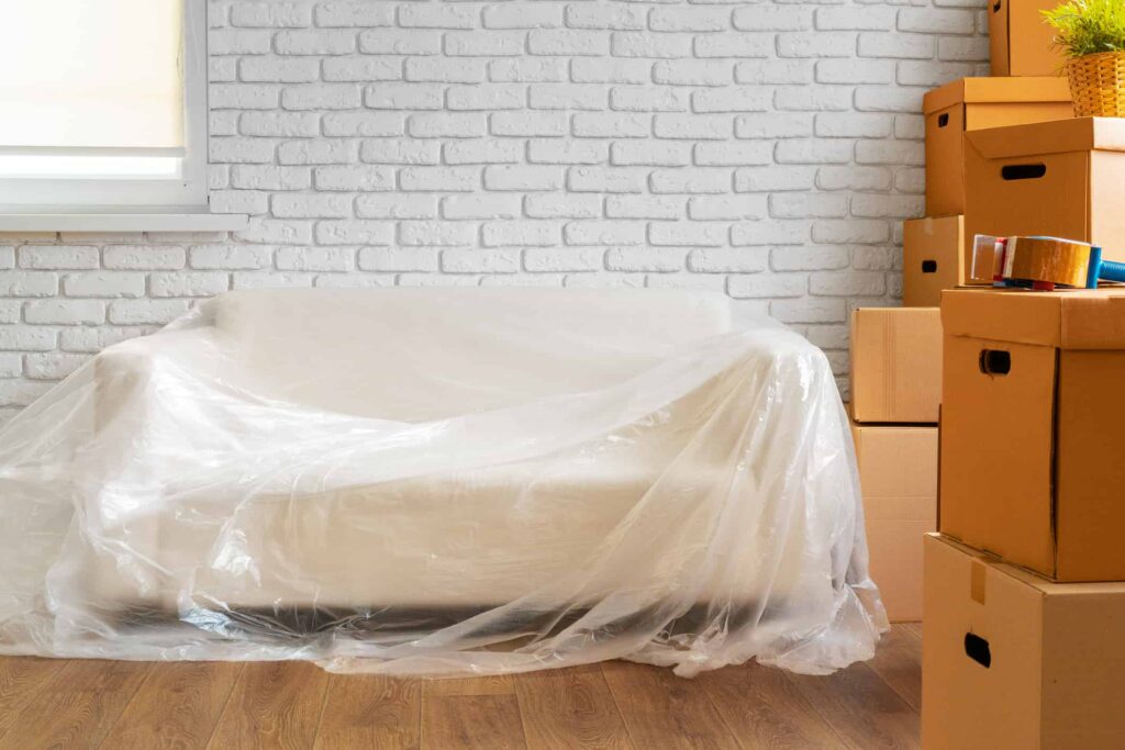 A white couch sits wrapped in plastic next to several cardboard boxes.