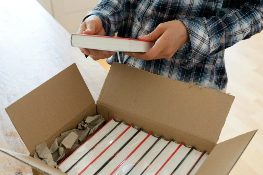 A man wearing a blue plaid shirt holds a book over a cardboard box neatly packed with more books