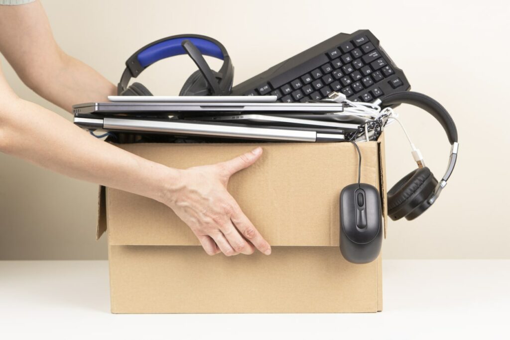 Hands hold a cardboard box full of assorted electronic devices: laptops, keyboard, headphones, and cords.