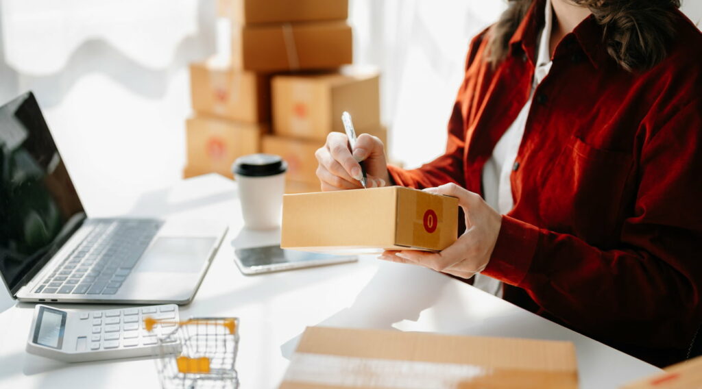 Person sits at a desk labeling cardboard boxes and using a laptop.