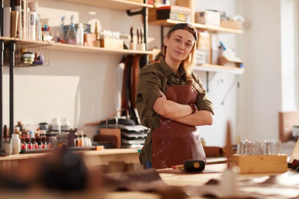 Smiling artisan wearing an apron stands with arms crossed in a workshop.