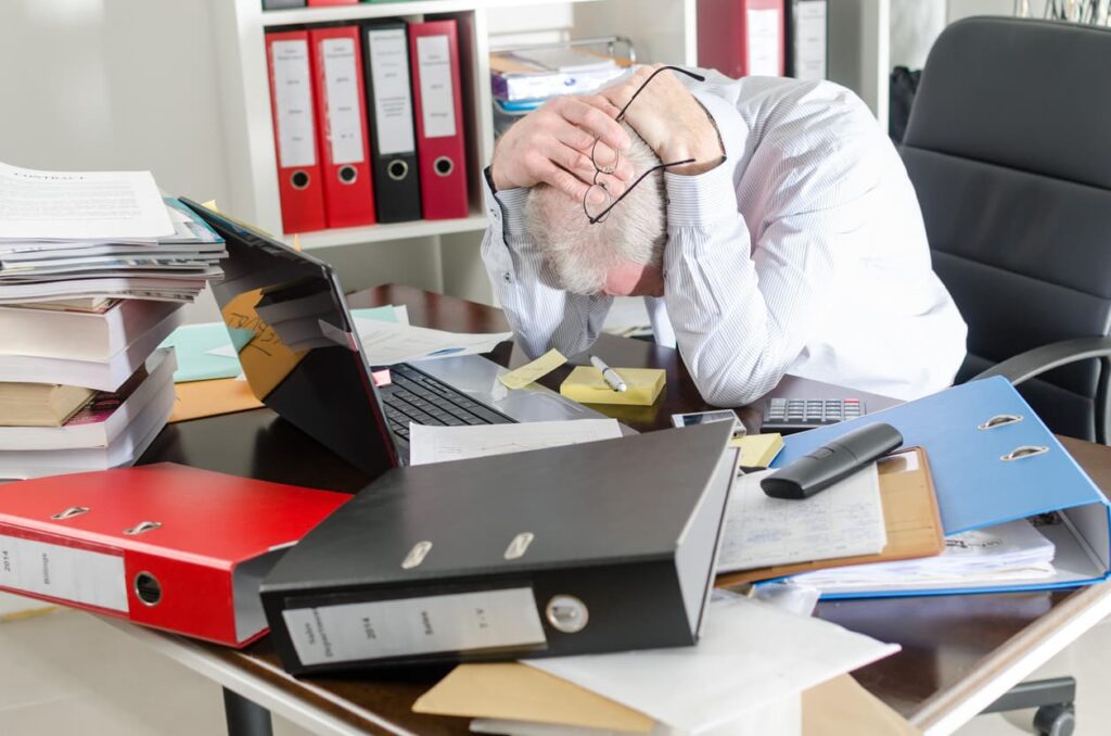 Stressed office worker hunches over messy desk with head in hands.