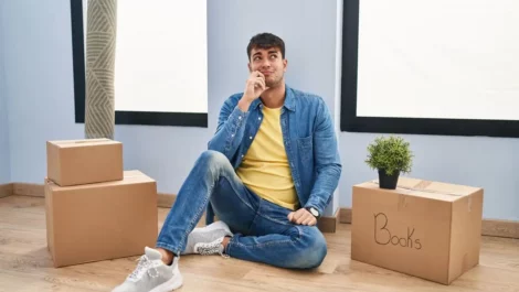 Person sits on floor next to packed moving boxes, contemplating.