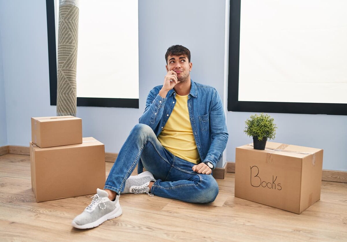 Person sits on floor next to packed moving boxes, contemplating.