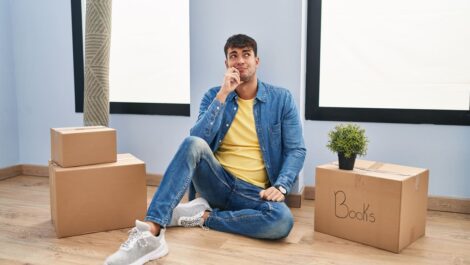 Person sits on floor next to packed moving boxes, contemplating.