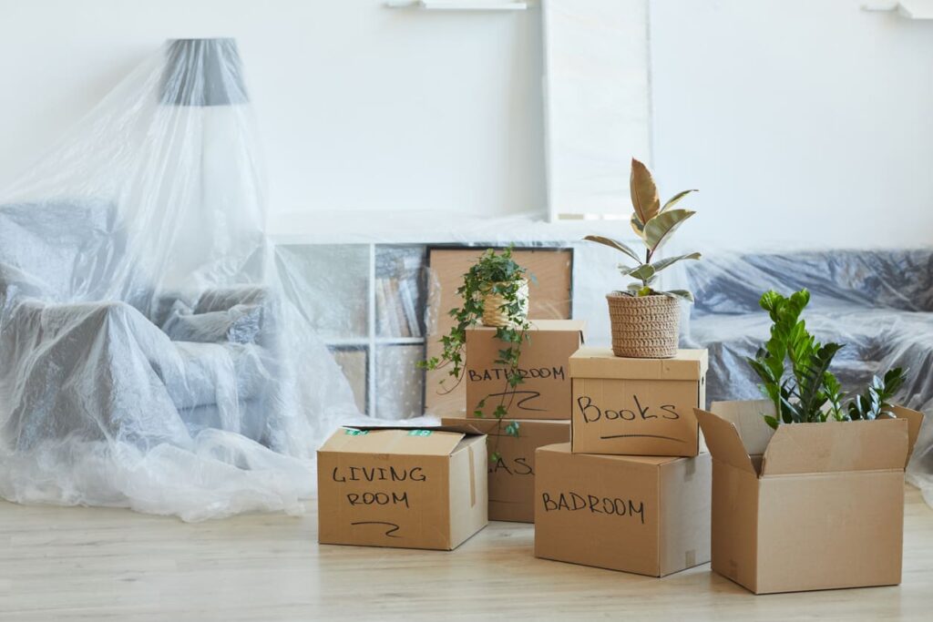 Moving boxes stacked up in a room with furniture covered by plastic.