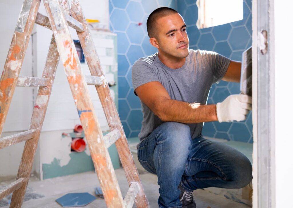 A man renovating bathroom tiles and crouching next to a paint-splattered ladder.