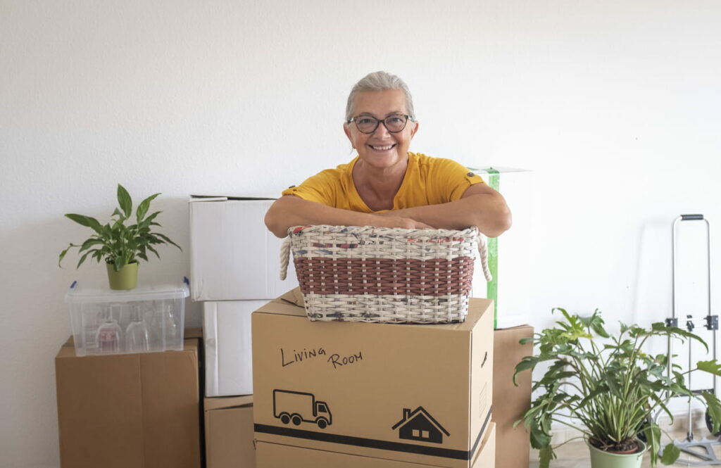 Portrait of a smiling woman with gray hair standing in a room with plants and items packed for storage.
