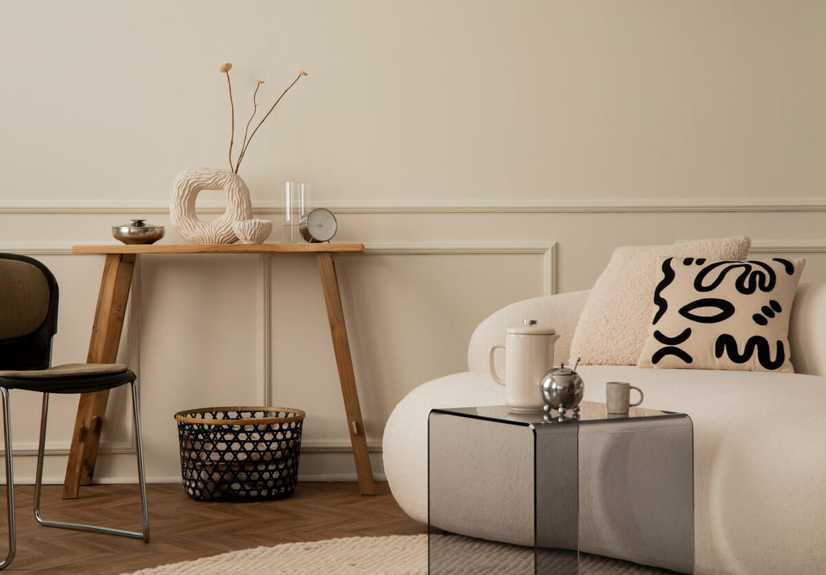 Cozy composition of living room interior with white sofa, glass coffee table, wooden bench, beige vase with dried flowers, patterned pillow, black chair, and accessories.