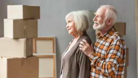 A senior couple stands together happily among several packed-up cardboard boxes.