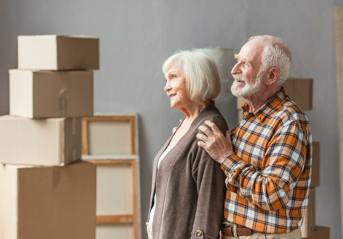 A senior couple stands together happily among several packed-up cardboard boxes.