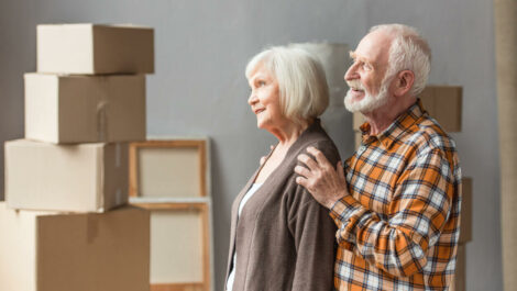 A senior couple stands together happily among several packed-up cardboard boxes.