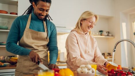 A couple smiles while chopping vegetables in a small apartment kitchen.