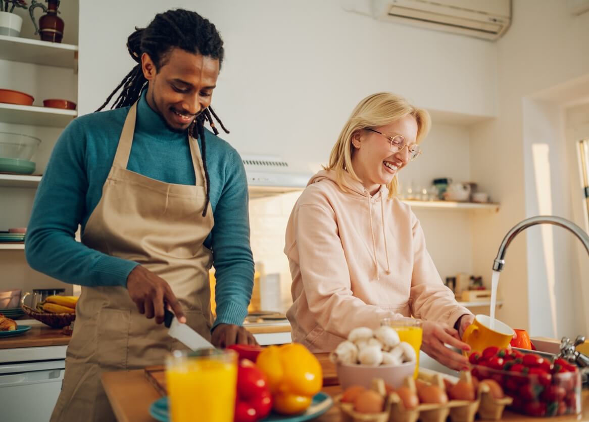 A couple smiles while chopping vegetables in a small apartment kitchen.