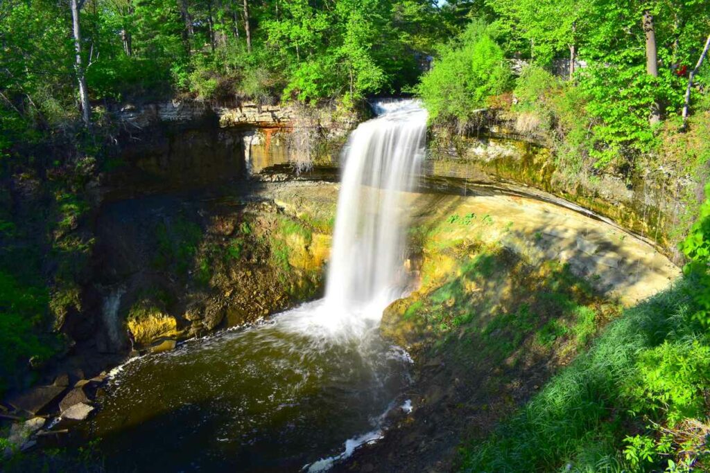 A waterfall, Minnehaha Falls, surrounded by rocks and green foliage during the day in Minneapolis, MN.