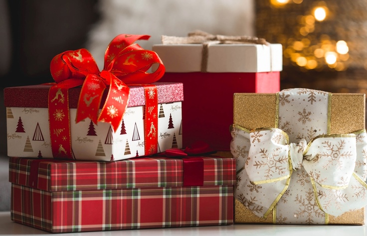 A pile of wrapped Christmas presents before a lit Christmas tree.