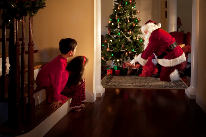 Two spying children peek at Santa Claus placing presents under their Christmas tree. 