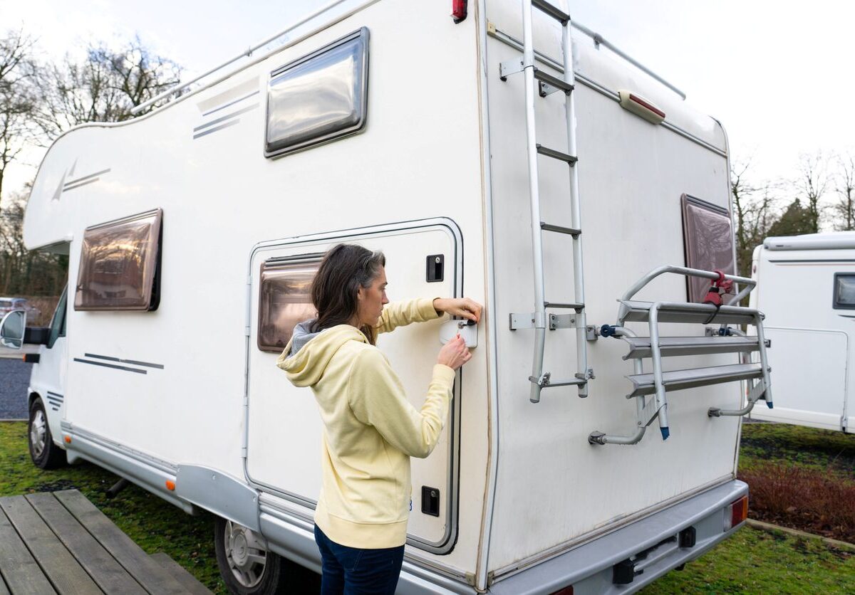 A woman in a yellow hoodie locks the back door of a white RV parked outdoors.