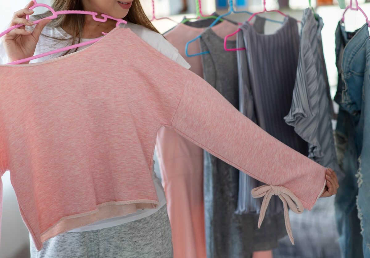 A woman holding a pink long-sleeve top while browsing spring clothing on hangers, including dresses and denim jackets.