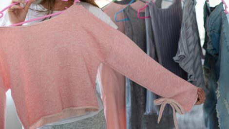 A woman holding a pink long-sleeve top while browsing spring clothing on hangers, including dresses and denim jackets.