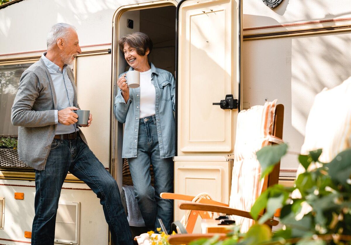 An older couple enjoying coffee together outside their RV on a sunny day, smiling and relaxing by the camper door.