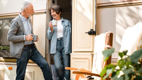 An older couple enjoying coffee together outside their RV on a sunny day, smiling and relaxing by the camper door.