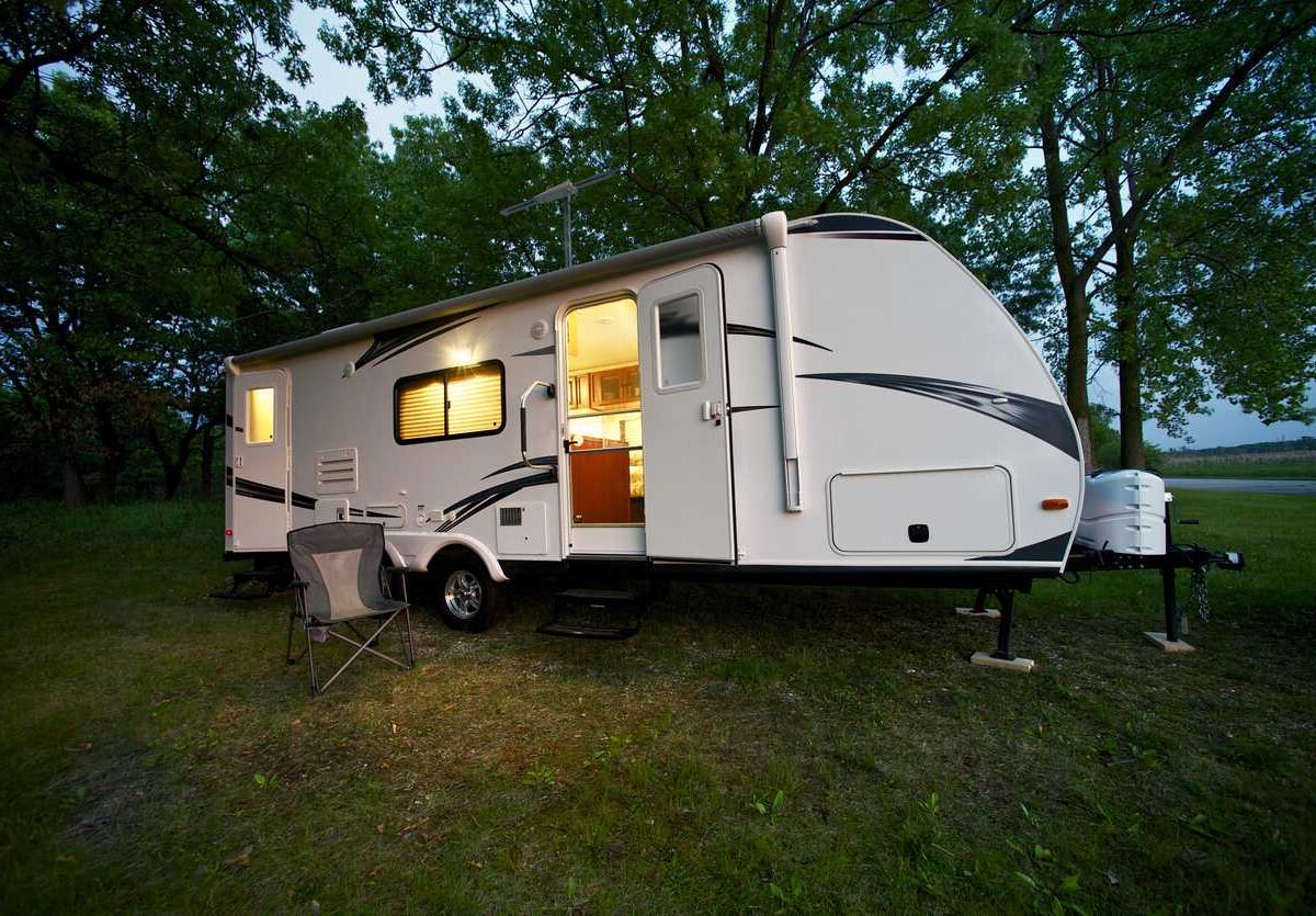 A white travel trailer parked at a campground, with lights on inside and a camping chair outside