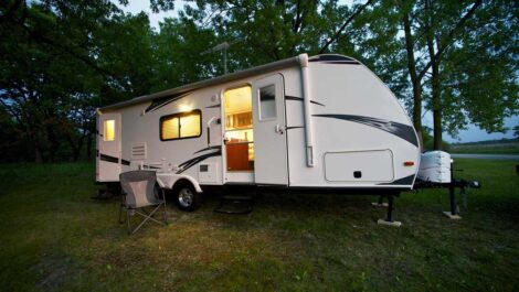 A white travel trailer parked at a campground, with lights on inside and a camping chair outside